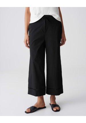 Femme portant un pantalon court noir ample avec une taille à cordon de serrage et des sandales noires à enfiler sur fond blanc.