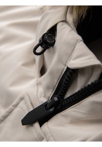 Fermeture éclair noire et bouton sur tissu beige, mettant en avant les détails de fixation d’une poche de veste avec couture texturée.