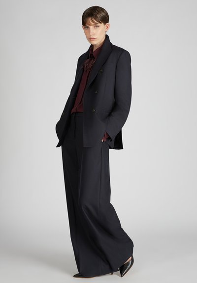 Blazer navy croisé à double boutonnage avec deux boutons, associé à un chemisier bordeaux foncé et un pantalon large, complété par des escarpins noirs pointus.