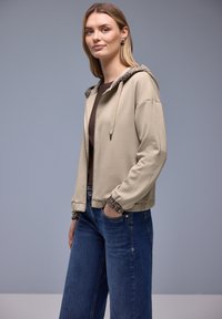 Beige jakke med lynlås, tekstureret hætte og manchetter. Glat stof, afslappet pasform, kombineret med blå jeans med brede ben for et afslappet look.