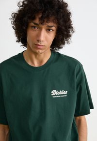Mørkegrøn bomulds t-shirt med korteærmer, med et hvidt logo på brystet. Enkel design, glat tekstur og afslappet pasform.