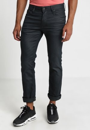 Mann trägt schwarze Slim-Fit-Jeans, an den Knöcheln hochgekrempelt, kombiniert mit schwarz-weißen Sneakers und einem korallenfarbigen Hemd.