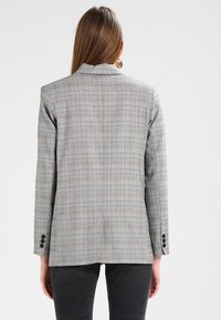 Blazer a scacchi grigio con un design strutturato, revers a punta e tre bottoni neri su ciascun polsino. Tessuto liscio con una vestibilità sartoriale.