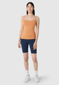 Camiseta sin mangas ajustada de color naranja con tirantes finos, combinada con pantalones cortos de ciclismo azul marino. Modelo lleva zapatillas blancas y calcetines de tobillo con estampado.