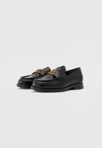 Schwarze Lederloafers mit glänzender Oberfläche, strukturiertem Material, runder Zehenpartie und goldfarbener Hardware-Akzent an der Vorderseite.