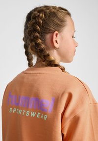 Pomarańczowy sweter z żeberkową fakturą. Na plecach znajduje się logo "hummel" w kolorze purpurowym oraz "SPORTSWEAR" w miętowym zielonym. Luźny krój.