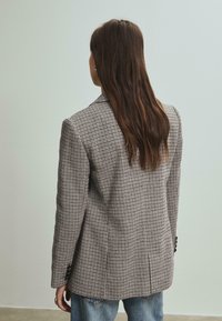 Blazer à motif pied-de-poule gris et marron avec une coupe structurée, revers crantés et deux boutons, porté avec un jean bleu délavé.