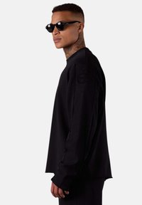 Sweat-shirt noir surdimensionné en tissu doux, avec un col rond et des manches longues dotées de détails cousus. Porté avec des lunettes de soleil noires.