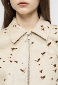 Jeune personne portant une veste crème avec des taches marron et une fermeture éclair argentée sur le devant, visible du cou à la poitrine.