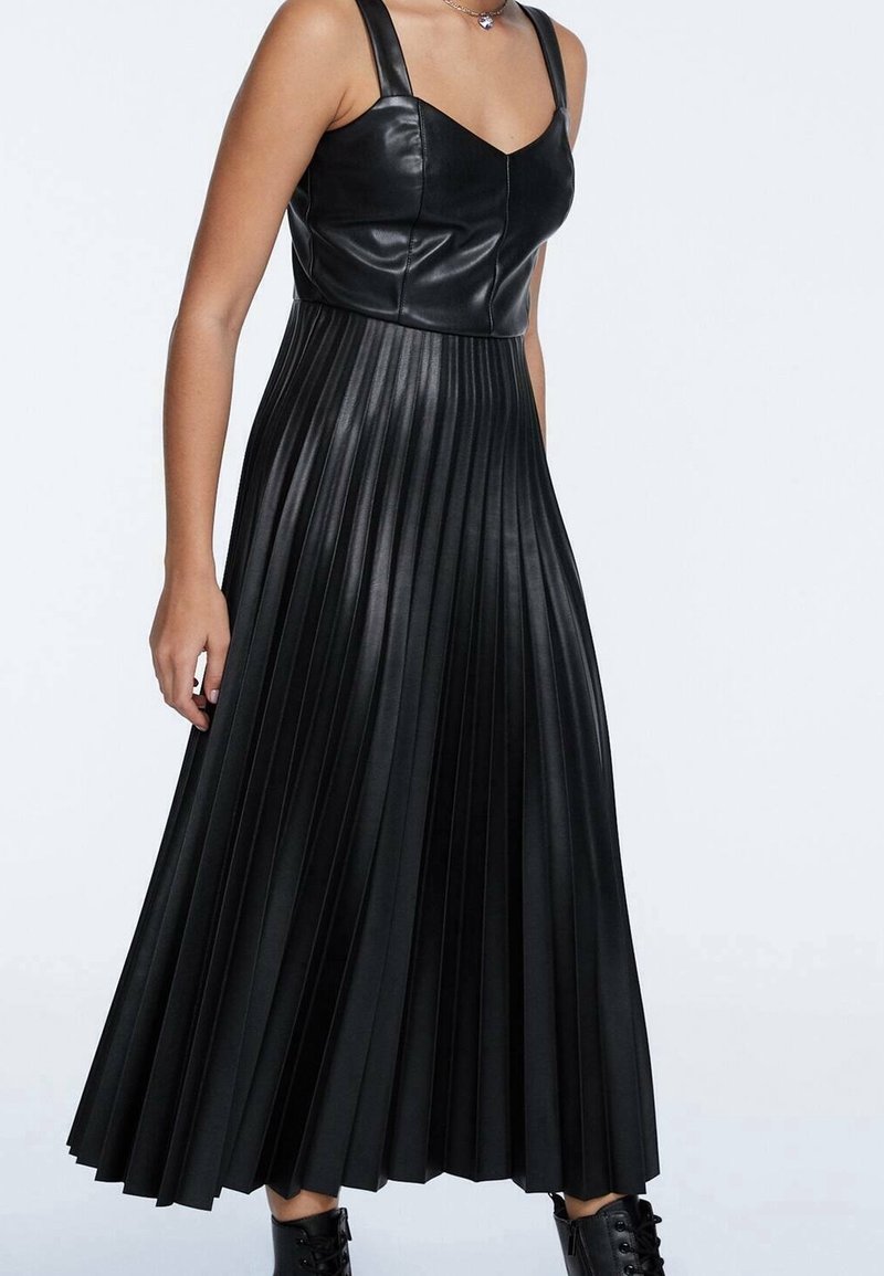 Femme portant un haut sans manches en similicuir noir et une jupe midi plissée assortie, avec des bottes noires lacées.