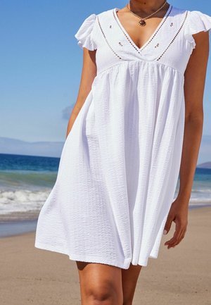 Robe en coton blanche avec des manches courtes à volants, décolleté en V et accents d broderie à l'avant. Coupe ample et tissu texturé, portée à la plage.