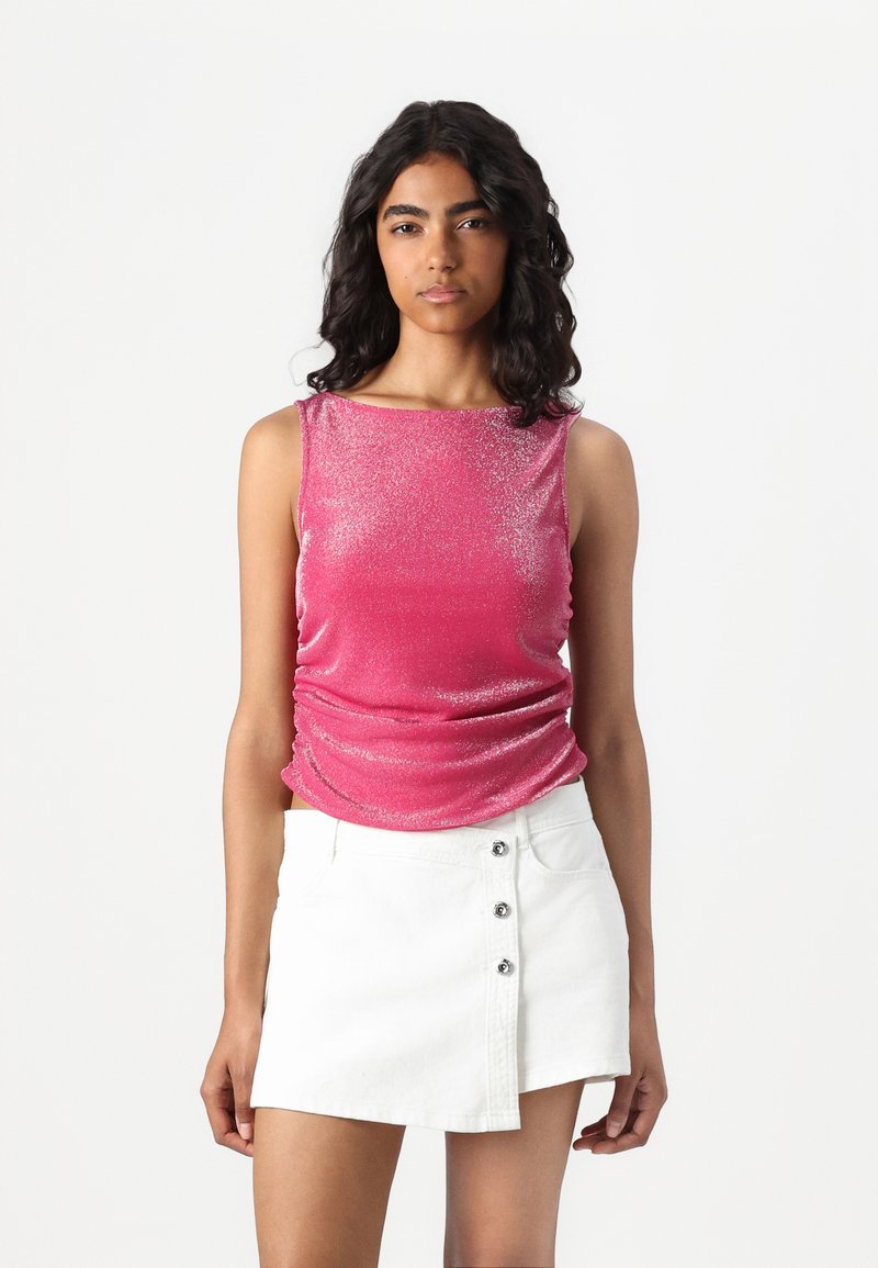 Rosa ermeløs topp med en blank tekstur, innsamlede sider, parret med et hvitt denim mini-skjørt med frontknapper. Ensfarget hvit bakgrunn.