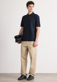 Camisa polo azul-marinho com colarinho e três botões, combinada com calças largas de tom bege claro e ténis pretos, segurando uma bolsa preta.