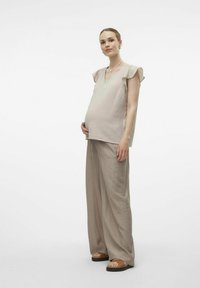 Ljusbeige graviditetsoutfit bestående av en kortärmad, strukturerad topp och löst sittande, vida byxor. Modellen står framför en neutral bakgrund.