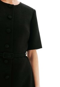 Robe noire en tissu texturé, col rond, manches courtes et six boutons assortis, maintenue par une ceinture.