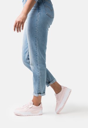 Zapatillas sintéticas rosas con un diseño elegante, suelas de goma blancas y detalles texturizados. Llevadas con vaqueros de mezclilla azul claro remangados.