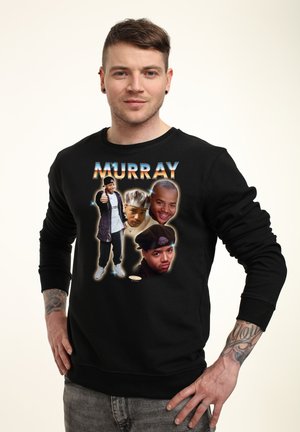 Homme avec des tatouages porte un sweat-shirt noir avec quatre portraits d’un homme et le texte "MURRAY" au-dessus sur un fond uni.