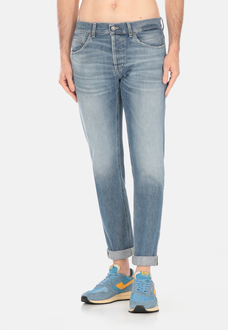 Jeans in denim azzurro chiaro con taglio slim, orli risvoltati e design a cinque tasche. Indossati con sneakers blu con dettagli arancioni e gialli.