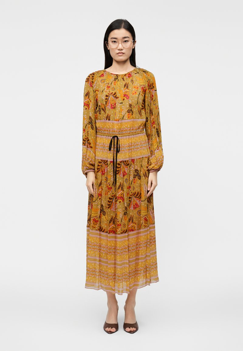 Μακρύ, κίτρινο φόρεμα με λουλούδια, μακριά μανίκια, ελαστική μέση και μαύρο κορδόνι. Διαθέτει πολλαπλά σχέδια, φούστα με στρώσεις και διαφανή υφή.