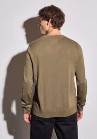 Olivgrön stickad tröja med rund hals och ribbade muddar. Slät textur med ett subtilt mönster och en avslappnad passform. Svarta byxor syns nedanför.