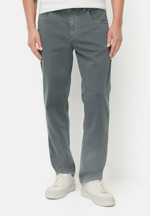 Mann trägt ausgewaschene blaue gerade geschnittene Hose und weiße Sneakers, steht vor einem einfachen weißen Hintergrund.