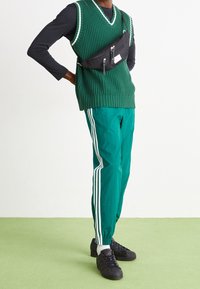 Grön ribbad stickad väst över en svart långärmad tröja, tillsammans med gröna träningsbyxor med vita ränder och svarta sneakers.