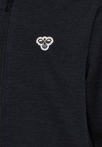Mörkgrå zip-up hoodie med en texturerad tyg, prydd med en liten broderad logotyp i vitt på det övre vänstra bröstområdet.