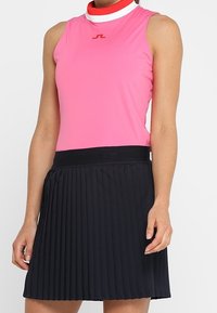 Růžový přiléhavý top bez rukávů s vysokým límcem, zdobený červenými a bílými detaily. Kombinován s černou plisovanou sukní s širokým páskem.