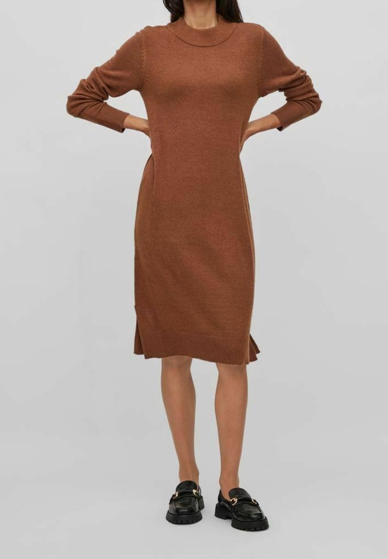 Femme portant une robe en maille marron à manches longues et des mocassins noirs, debout les mains sur les hanches sur un fond gris uni.