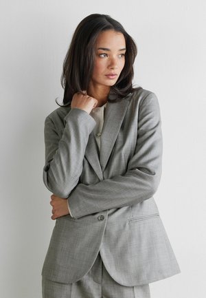 Frau mit langen dunklen Haaren, die einen grauen maßgeschneiderten Anzug trägt, steht vor einer schlichten weißen Wand und schaut mit nachdenklichem Ausdruck zur Seite.