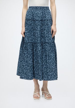 Frau trägt einen gestuften marineblauen Midirock mit Blumenmuster, elastischem Bund und weißen Riemchensandaletten mit Absatz.