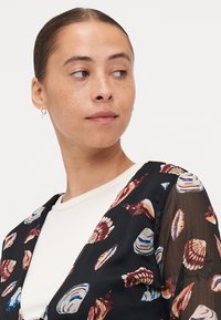 Blusa nera trasparente con motivo a conchiglia nei colori rosso, blu e bianco. Presenta un profondo scollo a V e maniche fluide sopra un top crema.