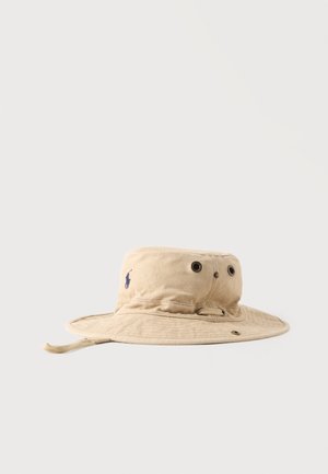 Béžový klobúk bucket s širokým okrajom, ventilačnými otvormi, nastaviteľným remienkom pod bradu a malým vyšívaným logom na boku.