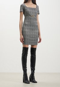 Vestido ajustado a cuadros en blanco y negro con escote cuadrado y mangas cortas, que presenta un acento de cadena dorada en la cintura. Combinado con botas brillantes negras hasta el muslo.