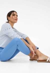 Gestreepte blouse met lange mouwen in blauw en wit, gecombineerd met lichtblauwe strakke broek en beige sandalen op een witte achtergrond.