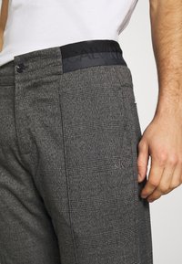 Grå rutiga byxor med en slät textur, som har en svart elastisk midja präglad med "CALVIN" och en liten "ck"-logotyp på sidan.