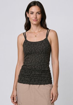 Frau mit dunklem Haar, die ein schwarzes Tanktop mit Leopardenmuster und einen beigen Rock trägt, steht vor einem schlichten hellen Hintergrund.
