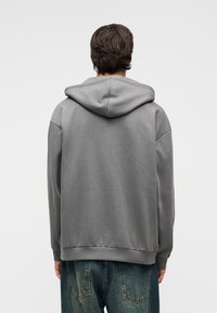 Άνδρας με πλάτη φορώντας μια φαρδιά γκρι ζακέτα με κουκούλα κατεβασμένη και υπερμεγέθη ξεθωριασμένα μπλε τζιν, μπροστά σε απλό φωτεινό φόντο.