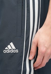 Jūriniai sportiniai kelnės su baltais šoninės juostelėmis, su išsiuvinėtu Adidas logotipu. Medžiaga lygi, sufunkcinė kišenė.