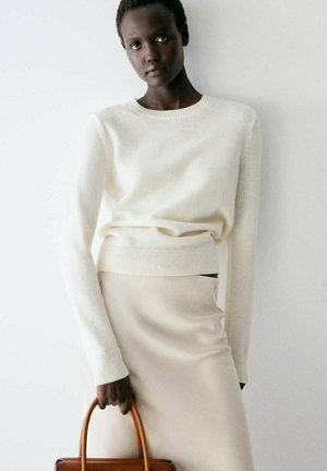 Mujer con suéter blanco y falda beige sosteniendo un bolso marrón contra un fondo claro y liso.