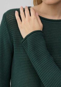 Grüner gestrickter Pullover mit einem strukturierten Muster, rundem Ausschnitt und gerippten Bündchen. Nahaufnahme einer Hand auf dem Schulterbereich zeigt das Detail des Stoffes.