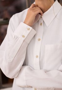 Personne portant une chemise blanche boutonnée avec une poche avant, touchant le cou d’une main et croisant l’autre bras.
