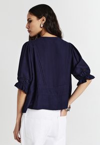 Donna vista da dietro che indossa una blusa blu marine con maniche a sbuffo e pantaloni bianchi, accessoriata con un orecchino d'argento e braccialetti.