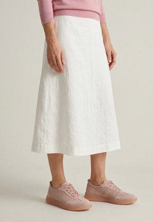 Žena ve bílé sukni po kolena, růžovém svetru a růžových teniskách s tkaničkami stojí před jednoduchým pozadím.