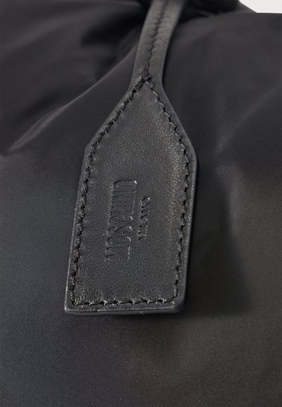 Κοντινό πλάνο μαύρης δερμάτινης ταμπέλας με ανάγλυφη την ένδειξη «HUGO SANTINO MILANO», ραμμένη πάνω σε μαύρο ύφασμα.