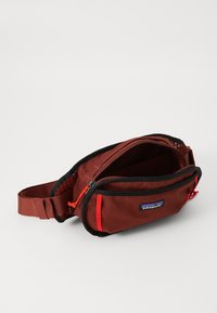 Sac en toile marron avec des accents noirs, doté d'un compartiment principal zippé, d'une tirette rouge et d'une sangle réglable. Logo Patagonia visible.