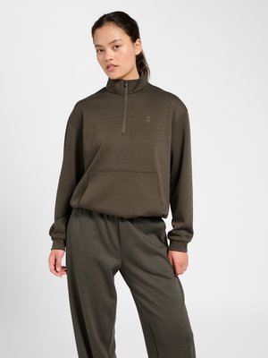 Femme portant un sweat-shirt vert olive à demi-fermeture éclair avec poche avant et pantalon ample assorti, posant devant un fond gris clair.
