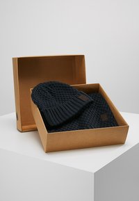 Set de căciulă și fular tricotat de culoare bleumarin în cutie din carton maroniu. Materialul are un model texturat, cu un accent din piele pe fiecare articol.