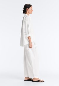 Femme debout de profil, portant un chemisier blanc ample, un pantalon blanc large et des tongs noires sur un fond blanc uni.