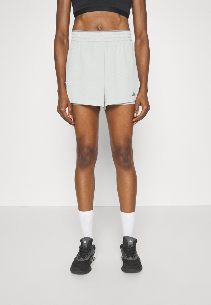 Pantaloni sportivi grigio chiaro con cintura elastica, tasche laterali e logo Adidas in basso a sinistra. Indossati con scarpe da ginnastica nere e calze bianche.